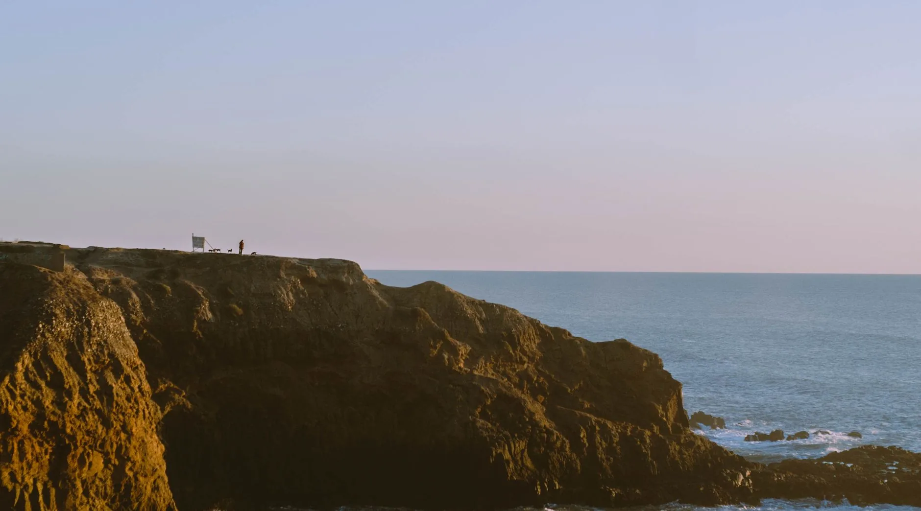A glass-walled house cantilevered over a cliff edge above the ocean at dusk
